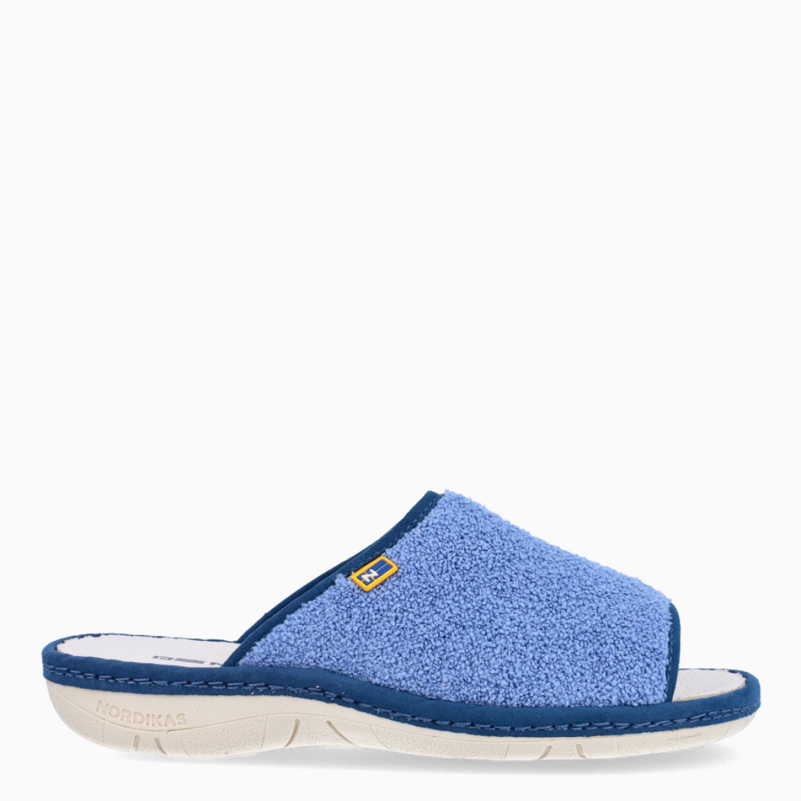 Zapatillas de casa de la marca Nordikas, modelo KAYA. Exterior de las zapatillas en toalla color azul. Interior de las zapatillas en piel troquelada color blanco. Zapatilla con suela plana. Estas zapatillas Nordikas KAYA son el complemento perfecto que garantiza comodidad y ligereza al estar por casa.