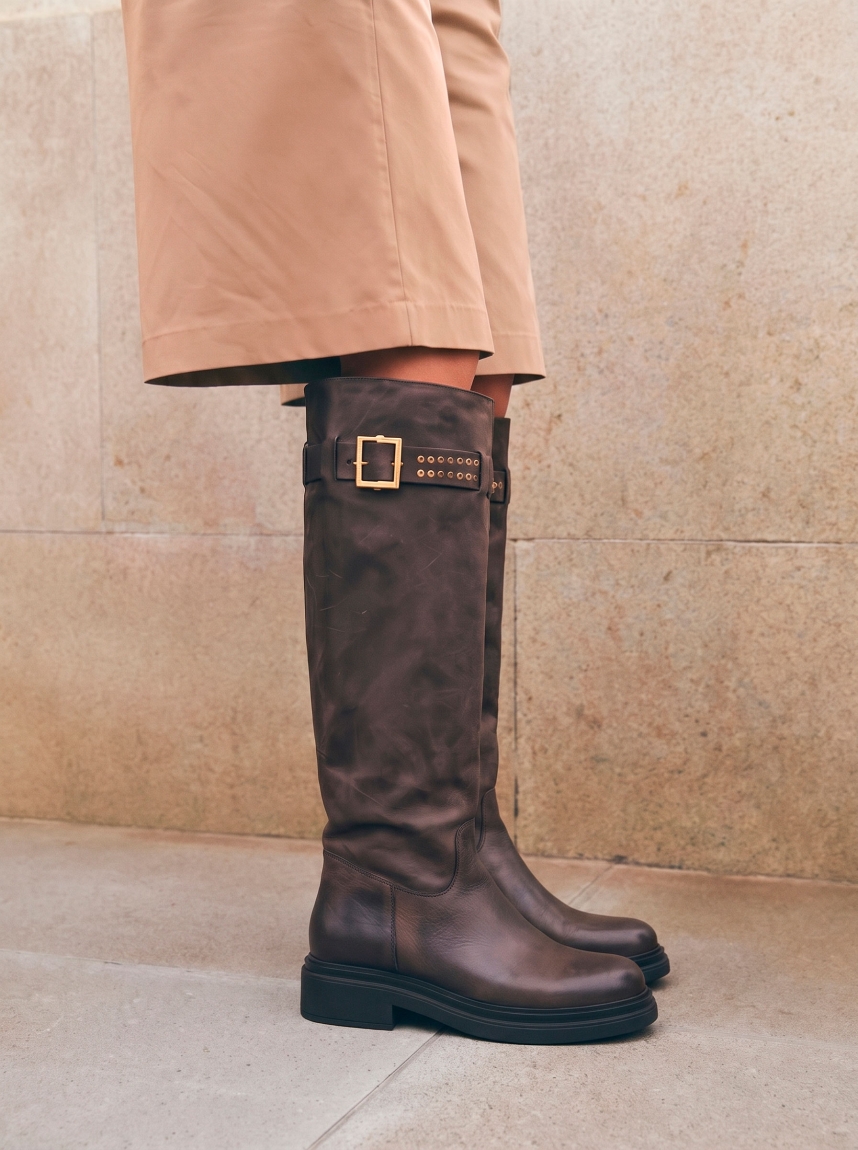 Botas altas de la marca Itse, modelo BALMA. Botas elaboradas en piel color taupe oscuro con hebilla y tachuelas en parte alta exterior. Botas sin cierre y con una punta redonda. Botas Itse con plataforma de 3cm de altura, que da una comodidad en cada paso. Botas Itse BALMA con un estilo moderno y contemporáneo, perfecto para usarlo con diferentes looks.