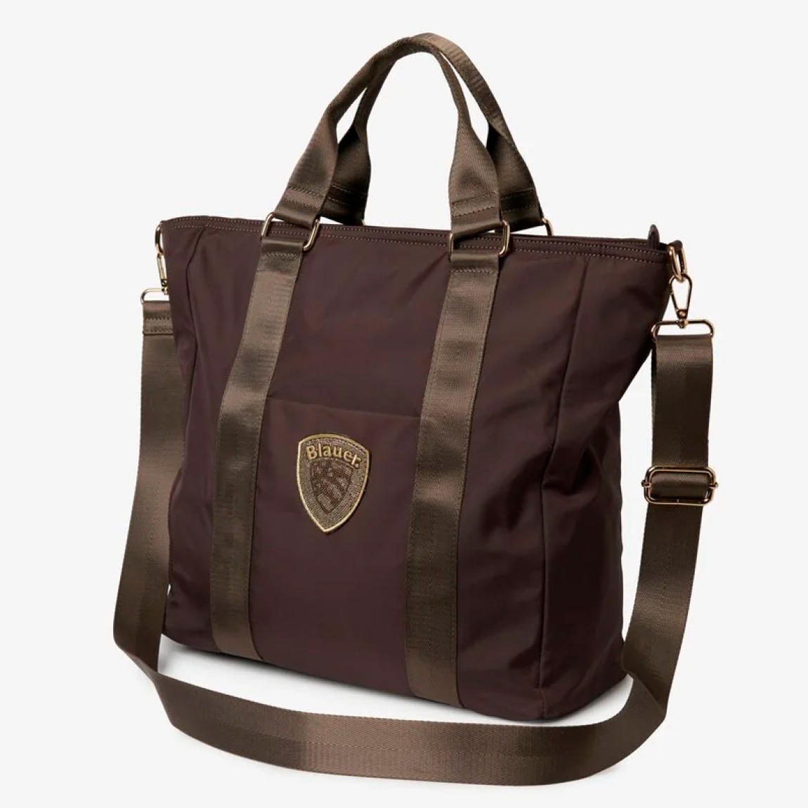 Bolso shopper Blauer, modelo SILVA01. Bolso fabricado en nailon técnico color marrón con detalles dorados. El diseño se caracteriza por amplias asas de cinta brillante, un bolsillo exterior plano con escudo del logo metalizado y una correa desmontable a juego. Este tote Blauer SILVA01 espacioso y ligero representa la combinación perfecta entre un estilo chic y funcionalidad para la mujer contemporánea. Medidas: 35 x 33 x 14cm