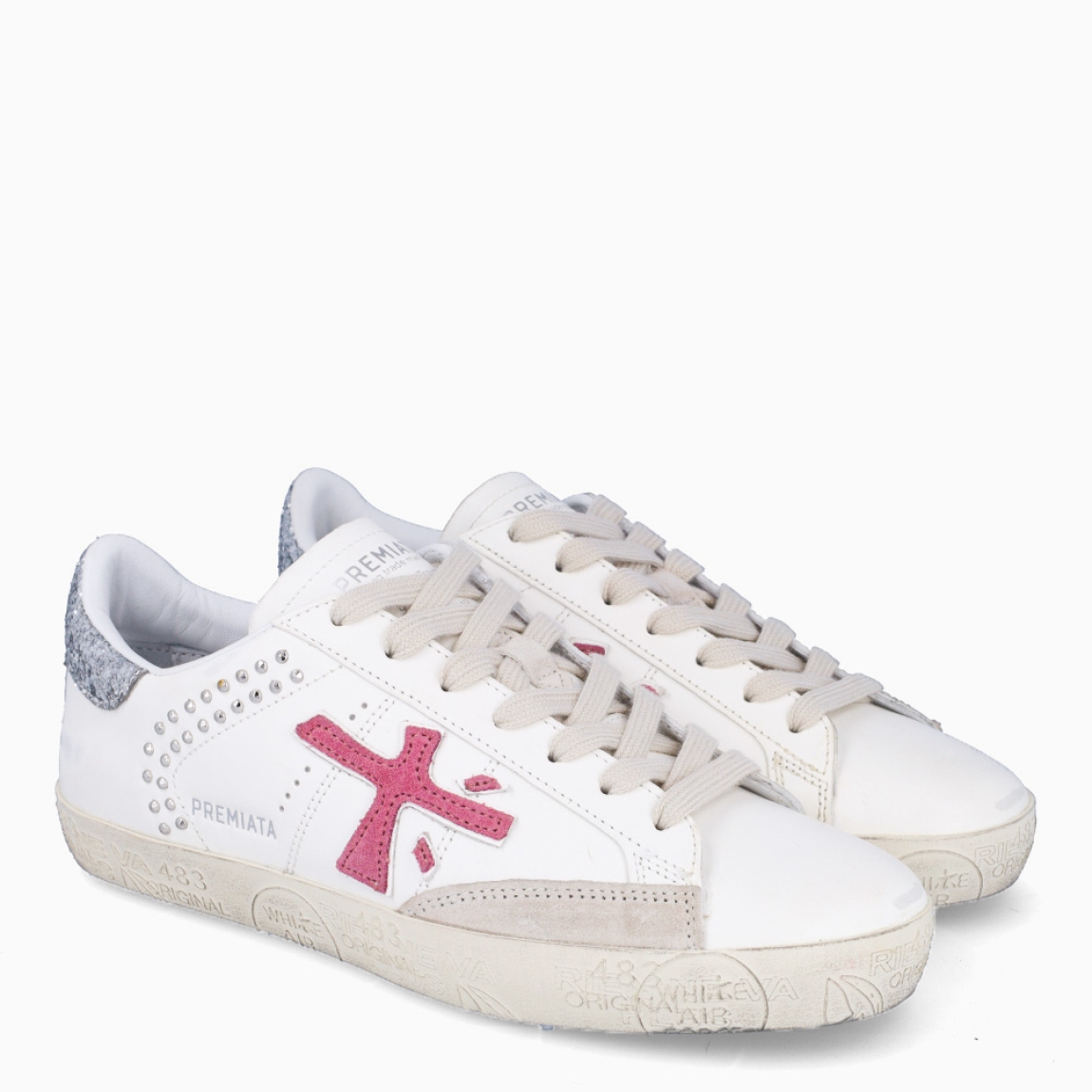 Deportivas de la marca Premiata, modelo STEVEND. Exterior de las deportivas en piel color blanco con detalle en tachuelas y glitter plateado en el talón. Logo de color fucsia. Interior de las deportivas en piel color blanco. Cierre de las deportivas con cordones blancos. Deportivas Premiata Stevend tienen el diseño perfecto para temporada, no puede faltar este modelo clásico en tu armario!