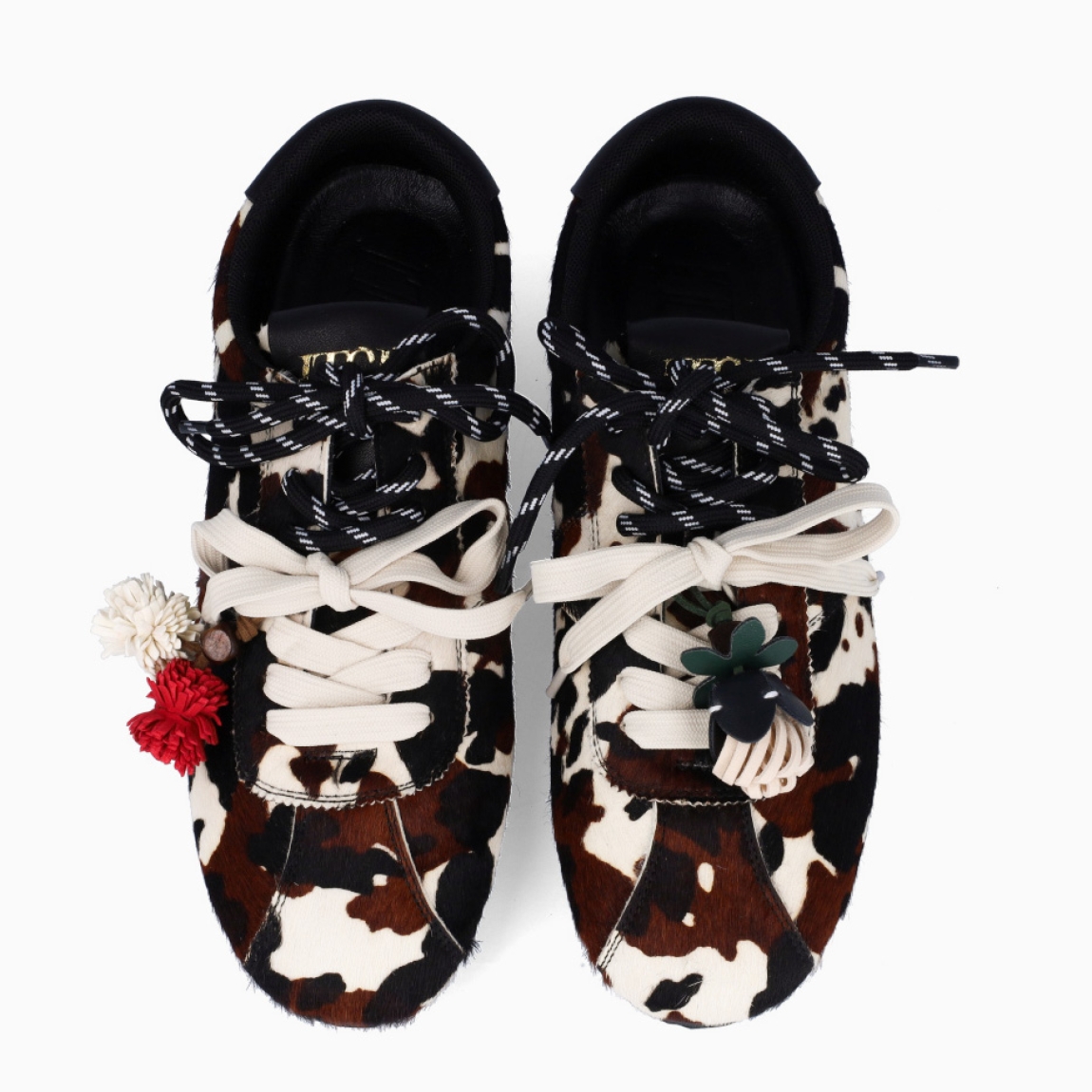 Deportivas de la marca Itse, modelo SALMA. Exterior en pelo con estampado animal print de vaca. Interior en textil color marrón. Cierre con cordones dobles, uno marrón y otro, beig. Deportivas Itse con tres charms sujetados a los cordones en forma de pompones y flor roja. Suela plana y de goma. Estas deportivas Itse Salma son un complemento básico y en tendencia para tus looks!