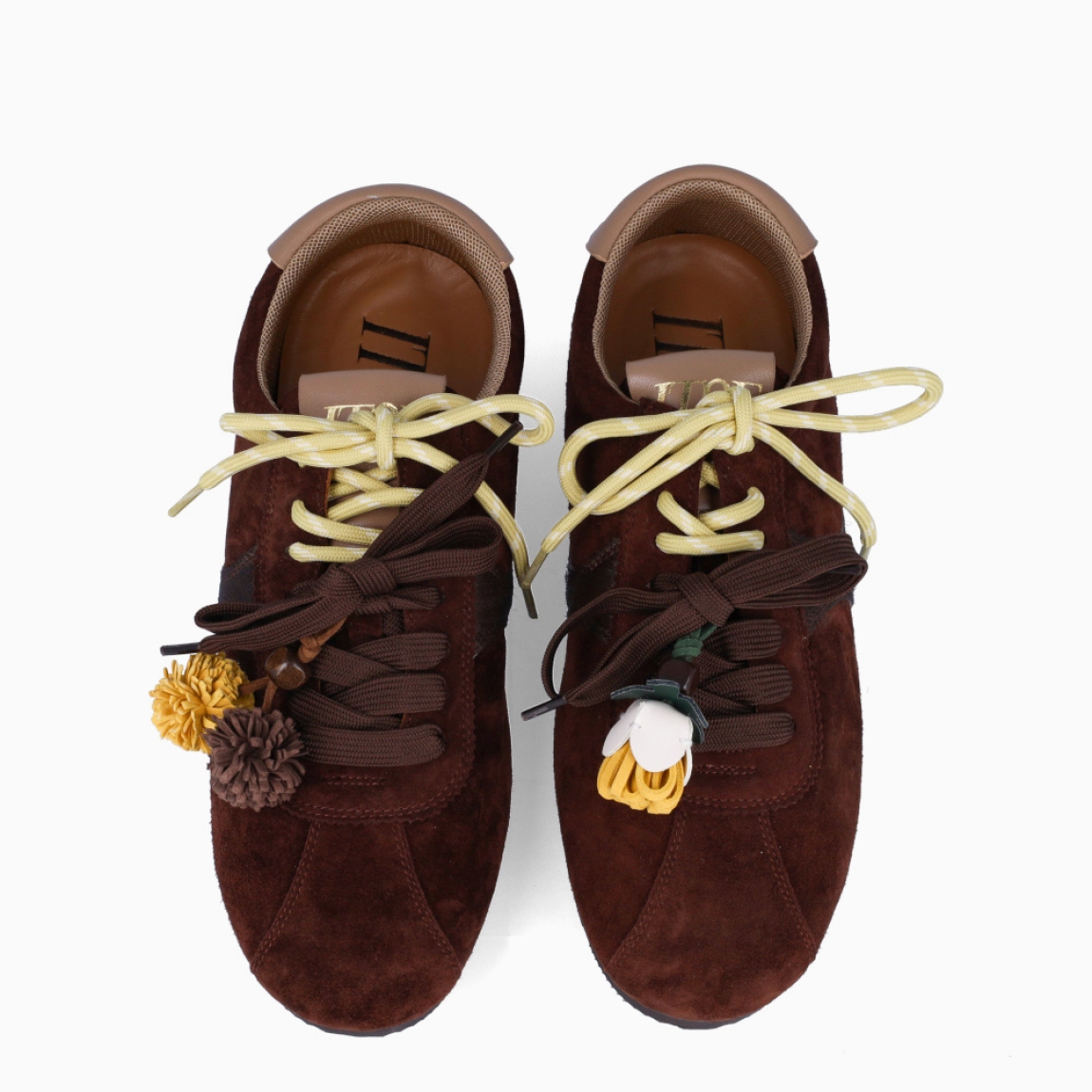 Deportivas de la marca Itse, modelo SALMA. Exterior en ante color marrón oscuro. Interior en textil color marrón. Cierre con cordones dobles, uno marrón y otro, amarillo. Deportivas Itse con tres charms sujetados a los cordones en forma de pompones y flor amarilla. Suela plana y de goma. Estas deportivas Itse Salma son un complemento básico y en tendencia para tus looks!