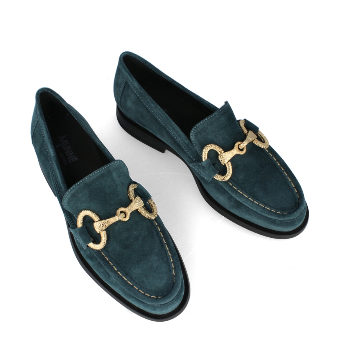 Mocasín de la marca Munné, modelo Indira. Exterior del mocasín de ante de color verde. Detalle de cadena dorada en la pala. Interior del mocasín de piel de color negro. Mocasín plano con plantilla acolchada para garantizar el máximo confort posible. Suela de goma con tacón bajo de 2 cm de altura. El mocasín de Munné, modelo Indira, es un mocasín clásico y cómodo, con un toque moderno debido a la cadena de adorno. Es el must-have de esta temporada, ya que queda bien con cualquier estilo y ocasión.