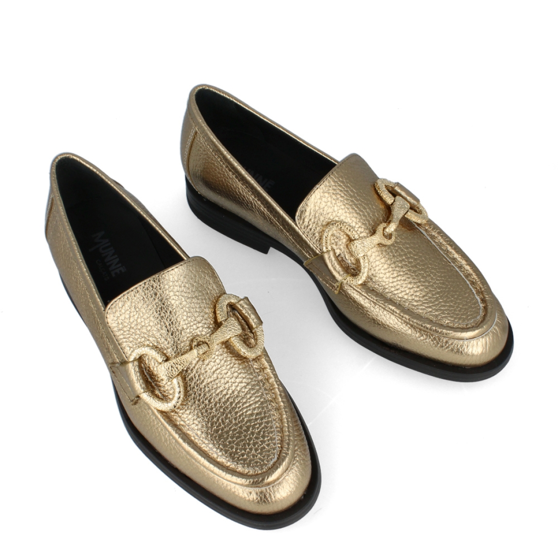 Mocasín dorado de la marca Munné, modelo Indira. Exterior del mocasín de piel metalizada de color dorado. Detalle de cadena dorada en la pala. Interior del mocasín de piel de color negro. Mocasín plano con plantilla acolchada para garantizar el máximo confort posible. Suela de goma con tacón bajo de 2 cm de altura. El mocasín de Munné, modelo Indira, es un mocasín clásico y cómodo, con un toque moderno debido a la cadena de adorno. Es el must-have de esta temporada, ya que queda bien con cualquier estilo y ocasión.