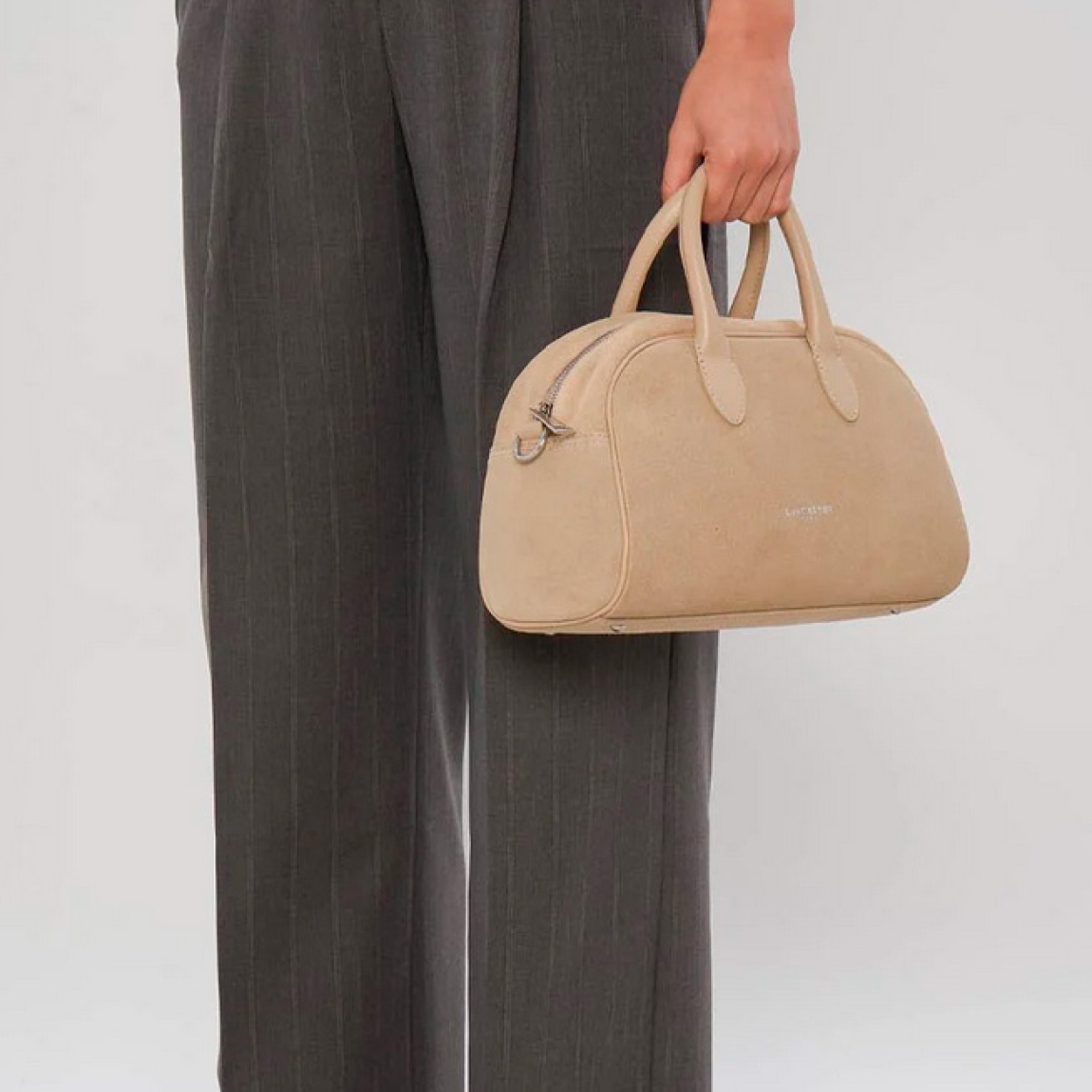 Bolso de la marca Lancaster, modelo BOWLING. Elaborado en ante color beig. Cierre con cremallera dos asas fijas. Además, de asas extraíbles para llevarlo cruzado. Cuenta con un bolsillo interior simple y un bolsillo interior con cremallera. Bolso Lancaster BOWLING perfecto para llevarlo a todos lado. Medidas: alto 18cm y ancho 29cm.