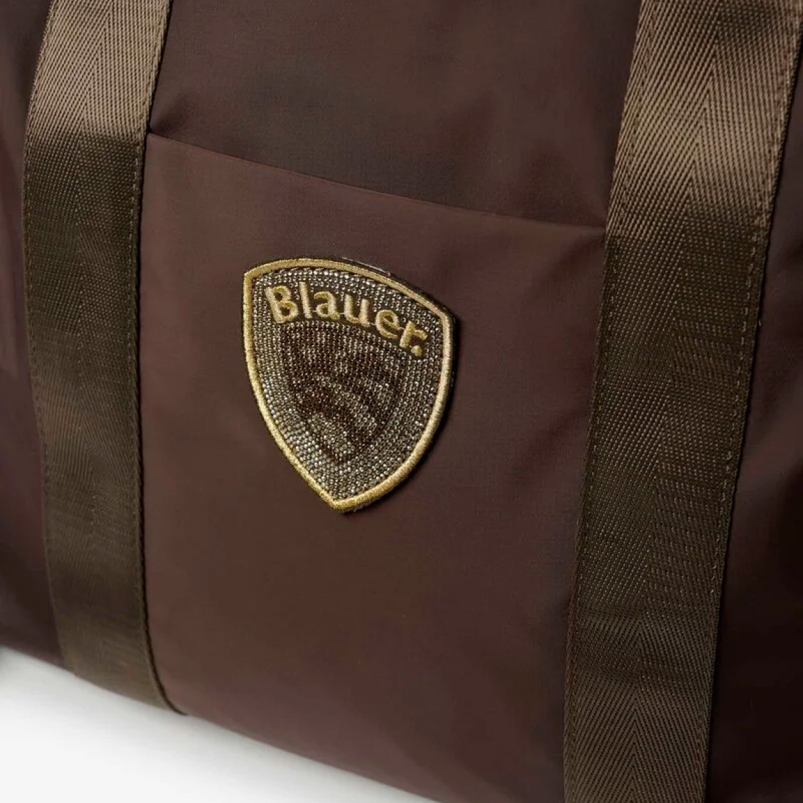 Bolso shopper Blauer, modelo SILVA01. Bolso fabricado en nailon técnico color marrón con detalles dorados. El diseño se caracteriza por amplias asas de cinta brillante, un bolsillo exterior plano con escudo del logo metalizado y una correa desmontable a juego. Este tote Blauer SILVA01 espacioso y ligero representa la combinación perfecta entre un estilo chic y funcionalidad para la mujer contemporánea. Medidas: 35 x 33 x 14cm