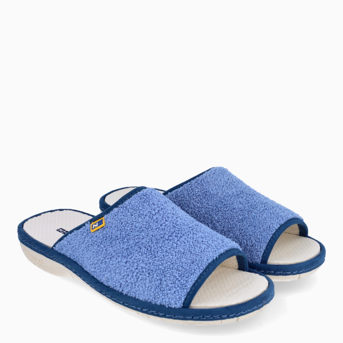 Zapatillas de casa de la marca Nordikas, modelo KAYA. Exterior de las zapatillas en toalla color azul. Interior de las zapatillas en piel troquelada color blanco. Zapatilla con suela plana. Estas zapatillas Nordikas KAYA son el complemento perfecto que garantiza comodidad y ligereza al estar por casa.