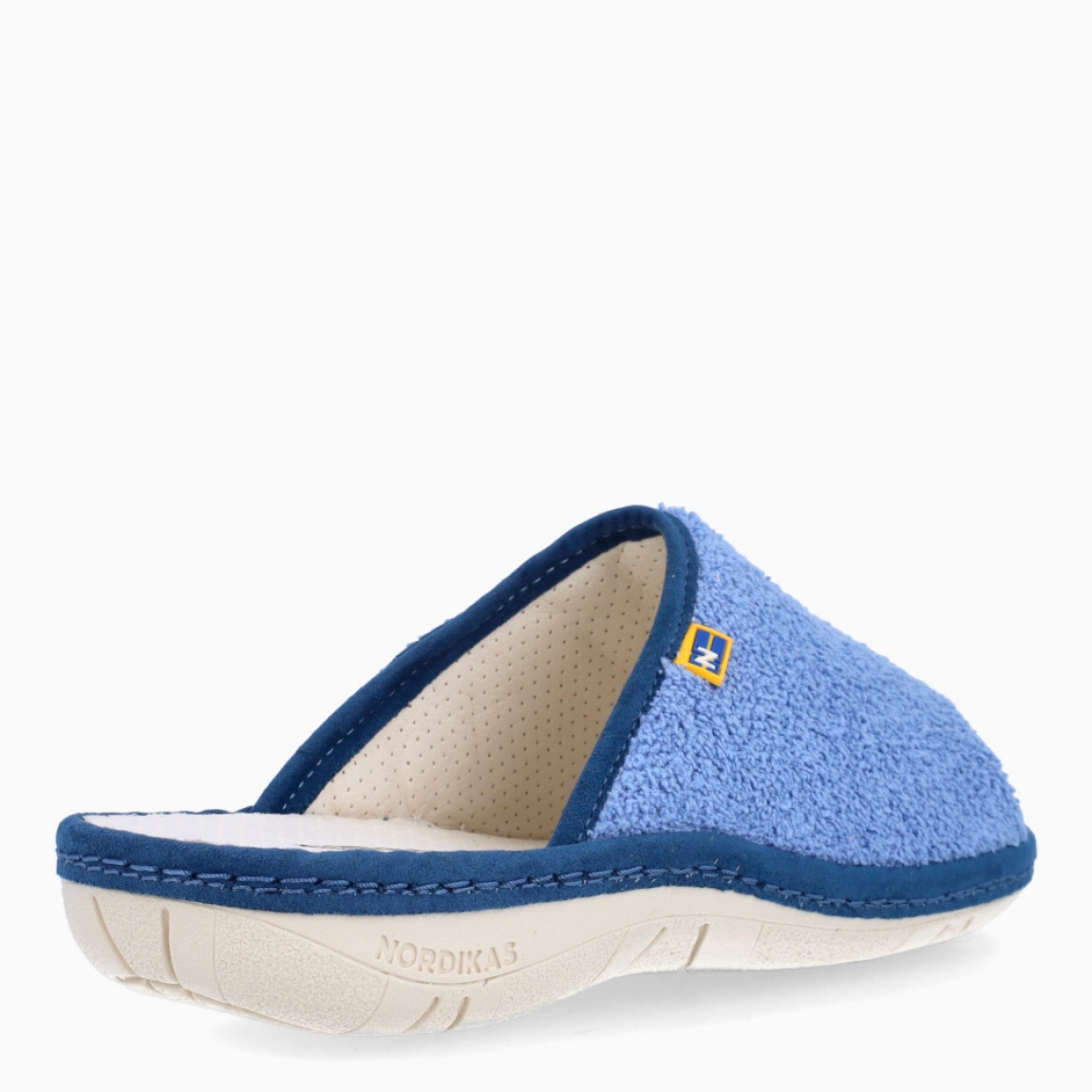 Zapatillas de casa de la marca Nordikas, modelo KAYA. Exterior de las zapatillas en toalla color azul. Interior de las zapatillas en piel troquelada color blanco. Zapatilla con suela plana. Estas zapatillas Nordikas KAYA son el complemento perfecto que garantiza comodidad y ligereza al estar por casa.