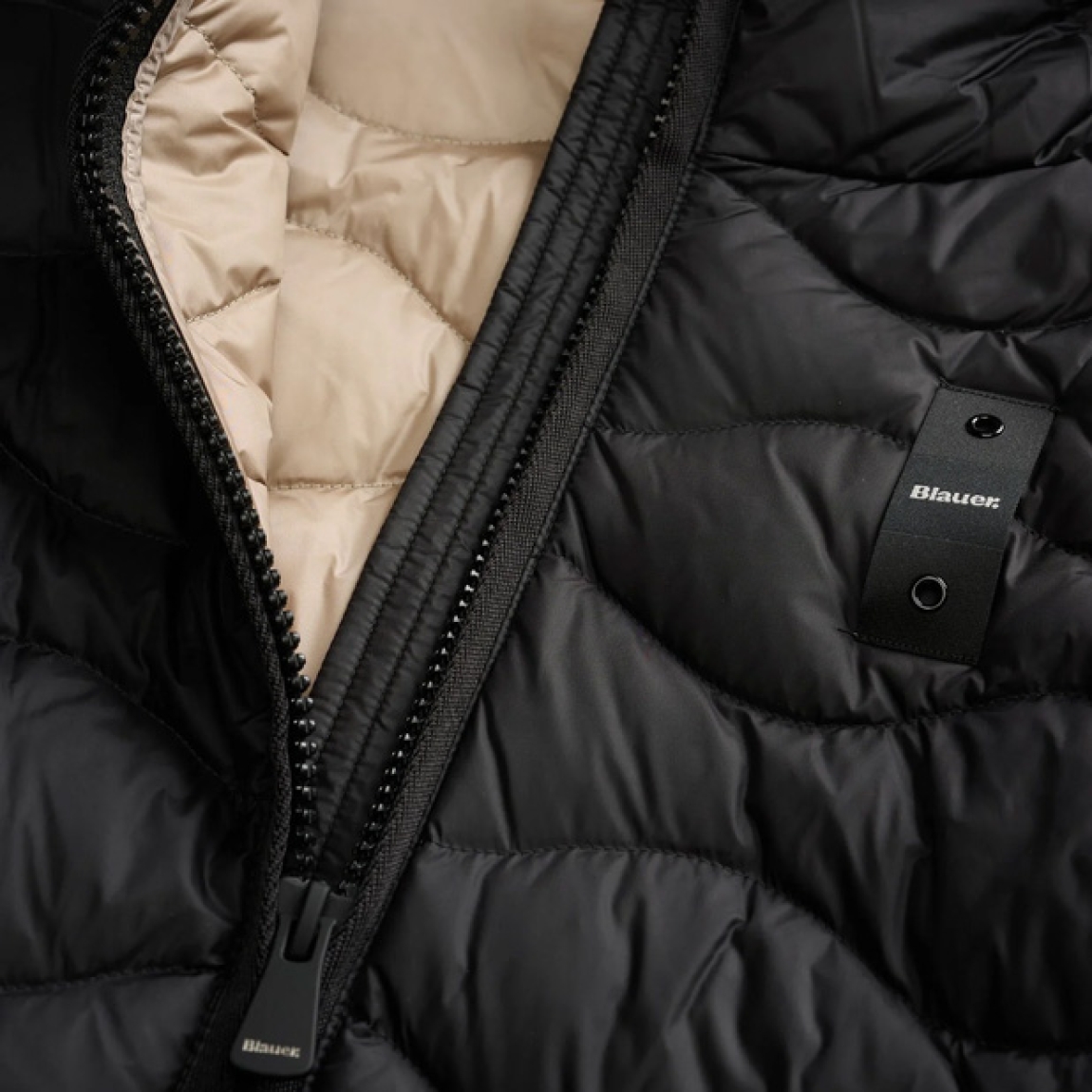 Chaqueta acolchada de la marca Blauer, modelo CAMELIA. Chaqueta con ondas horizontales de corte evasé. Esta prenda icónica de Blauer asegura un calor óptimo gracias al relleno natural de pluma (90/10 gr.165). Realizado en nylon brillante color negro con forro en contraste, capucha fija y elástico rayado, se completa con un refinado parche con purpurina. Chaqueta Blauer CAMELIA, ideal para esta temporada!