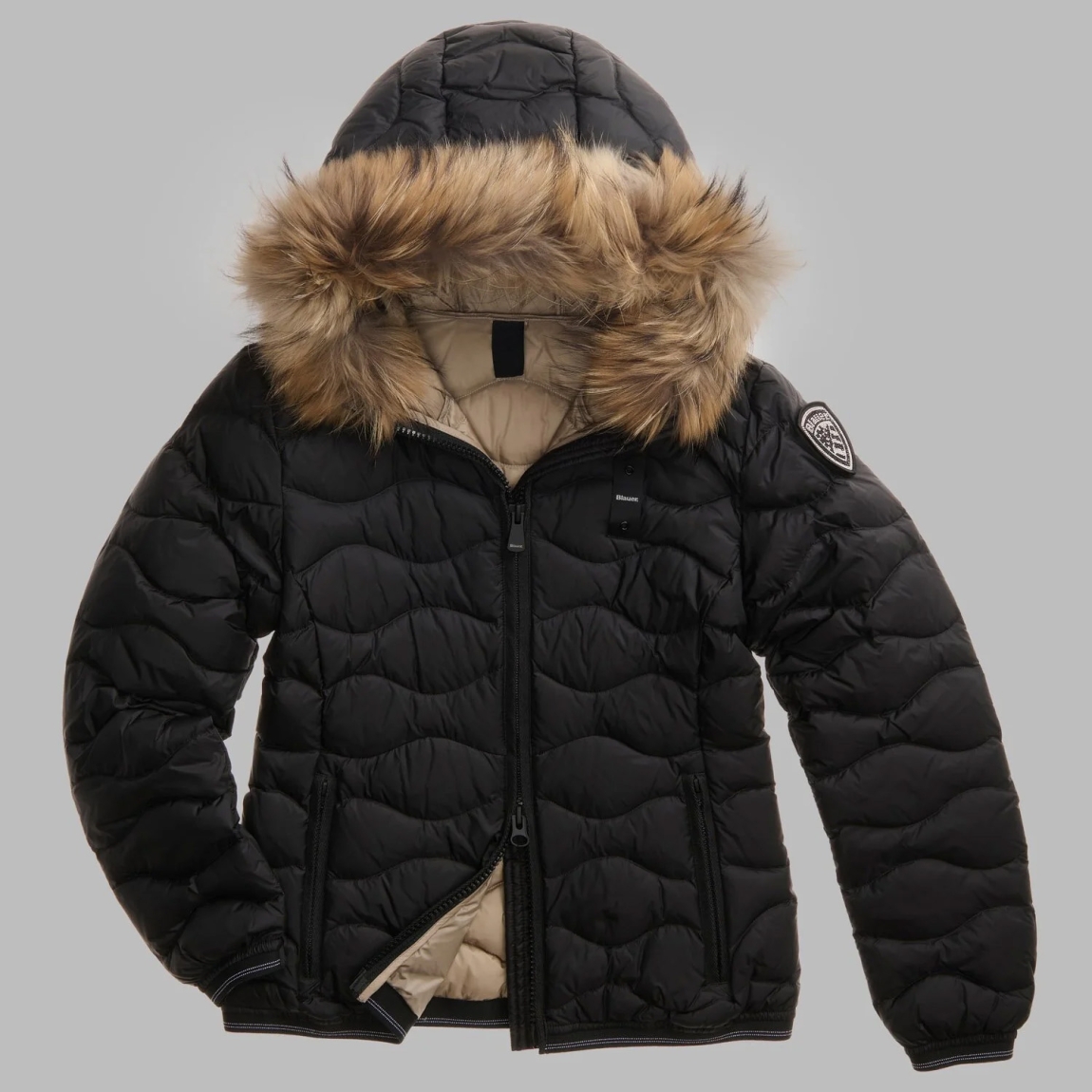 Chaqueta plumífera de la marca Blauer, modelo ALDIE. Chaqueta acolchada con ondas horizontales, embellecido con un borde de capucha en piel auténtica extraíble. Esta prenda icónica de Blauer ofrece un relleno natural (90/10 gr.175) cálido y no voluminoso. Realizado en nylon brillante color negro con forro en contraste, presenta un elegante parche con purpurina. Chaqueta Blauer ALDIE, ideal para los días fríos de esta temporada!