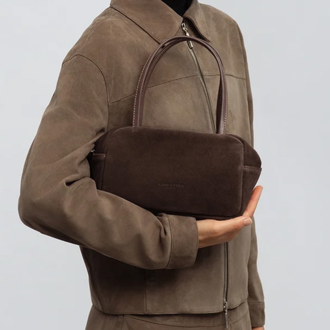 Bolso de mano de la marca Lancaster, modelo SUÈDE IT. Bolso confeccionado en piel de vacuno efecto terciopelo color marrón oscuro, ofreciendo un tacto suave y un specto elegante. Cuenta con un bolsillo trasero con cremallera, un bolsillo interior con cremallera y un bolsillo sencillo para organizar tus esenciales. Su cierre con cremallera garantiza seguridad y practicidad en el día a día. Bolso Lancaster equipado con una bandolera extraíble y ajustable, se lleva principalmente en la mano y también puede llevarse al hombro o cruzado. Medidas: alto 15cm y ancho 25cm.