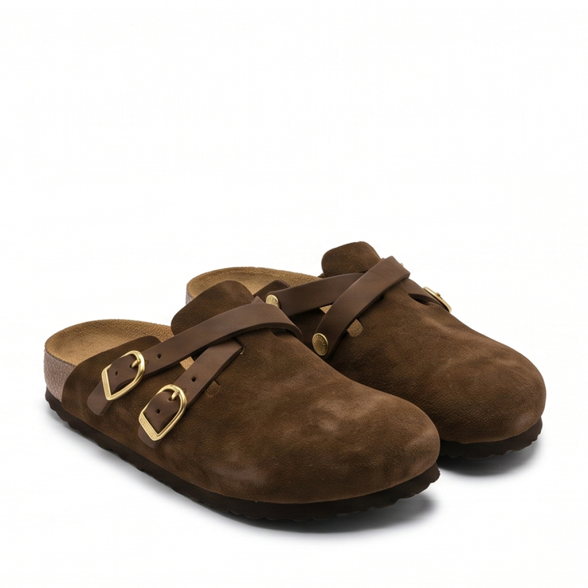 Zuecos de la marca Birkenstock, modelo BLAIR. Zuecos elaborados en piel de ante (serraje) color marrón chocolate, con detalle de tiras cruzadas en el empeine y elegantes hebillas ajustables en acabado dorado. Cuentan con la icónica plantilla anatómica de corcho y látex forrada en piel y suela plana de EVA ligera. Estos zuecos Birkenstock son ideales para complementar tus looks de temporada aunando tendencia y confort. No pueden faltar en tu armario.
