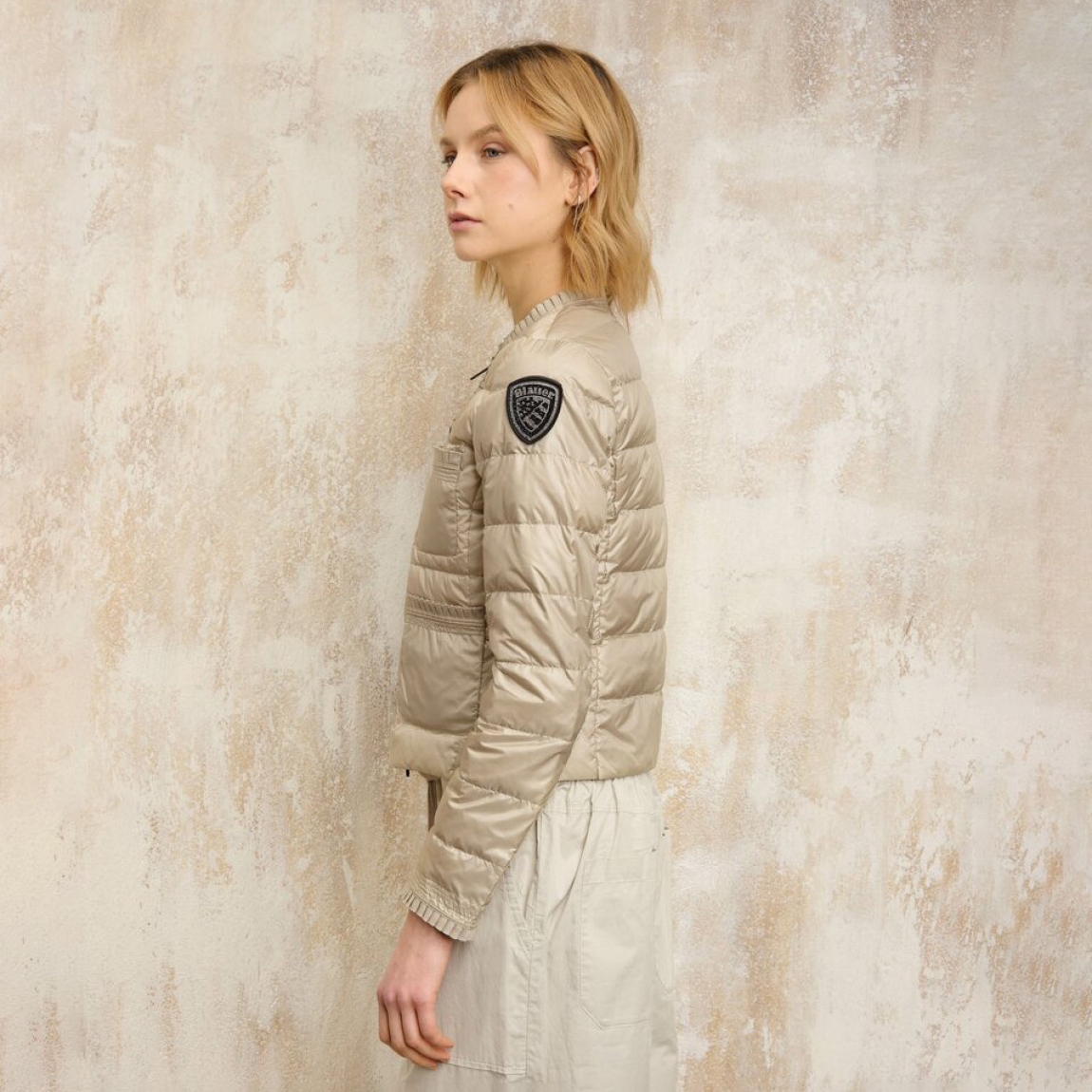 Chaqueta acolchada de la marca Blauer, modelo ROSE. Plumífero corto elaborado en naylon color beig. Chaqueta Blauer de cuello redondo con costuras en rayas horizontales. La chaqueta corta está adornada con una cinta plisada que le da a la prenda un estilo clásico y refinado. Cierre con cremallera. Acolchado con plumón 90/10  47 gr. Cuatro bolsillos. Bolsillo interior con velcro. Parche y pasante con logotipo. Chaqueta Blauer ideall para esta temporada!