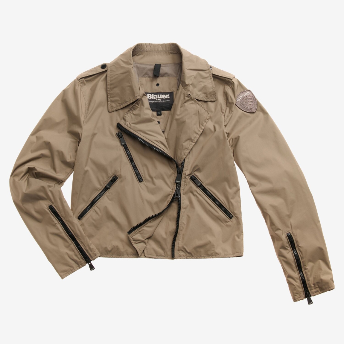 Chaqueta de cuero en nylon forrado color camel de la marca Blauer, modelo Zamora. Chaqueta con cuello de doble solapa para un look chic y desenfadado que quiere resaltar la figura. Cierre asimétrico izquierdo con cremallera. Bolsillos con cremallera y bolsillo pequeño con cremallera en el pecho. Bolsillos interiores con velcro. Parche con logotipo. Chaqueta Blauer Zamora ideal para esta temporada!