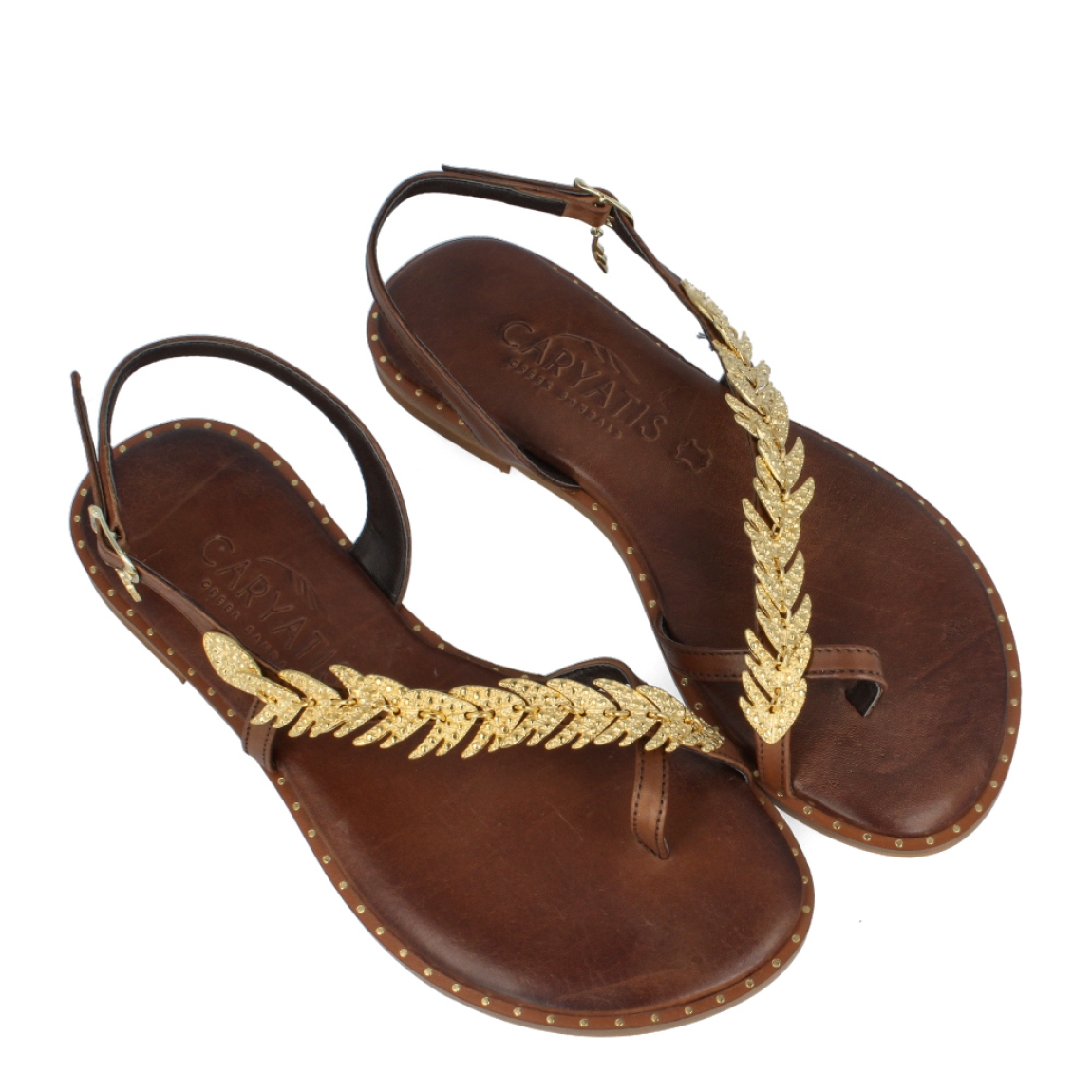 Esclavas de la marca Caryatis, modelo EIRA. Exterior de las esclavas en piel color marrón con adorno dorado metálico dorado en forma de hojas en la pala. Cierre de las esclavas con hebilla y suela plana. Estas esclavas Caryatis EIRA es un producto de alta calidad, pensado en la comodidad y elegancia. No puede faltar en tu armario de temporada.