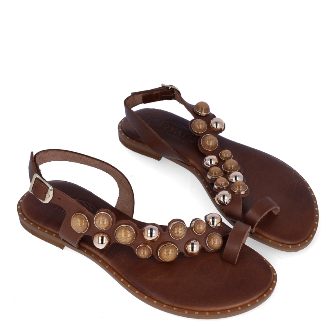 Esclavas de la marca Caryatis, modelo SENDRA. Exterior de las esclavas en piel color marrón con adornos en piedrería. Cierre de las esclavas con hebilla y suela plana. Estas esclavas Caryatis SENDRA es un producto de alta calidad, pensado en la comodidad y elegancia. No puede faltar en tu armario de temporada.