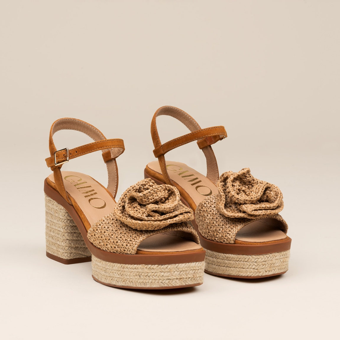 Sandalias de la marca Gaimo, modelo Luana. Elaboradas en rafia colo camel, combinan elegancia y comodidad con su propuesta en rafia natural, con un delicado adorno de flor y talón abierto con pulsera ajustable. Cierre con hebilla en tobillo y un tacón de 8cm de altura. Elaboradas artesanalmente en Arnedo, España, con materiales de alta calidad y 100% naturales, las sandalias Luana son la opción perfecta para quienes buscan elevar sus looks veraniegos.